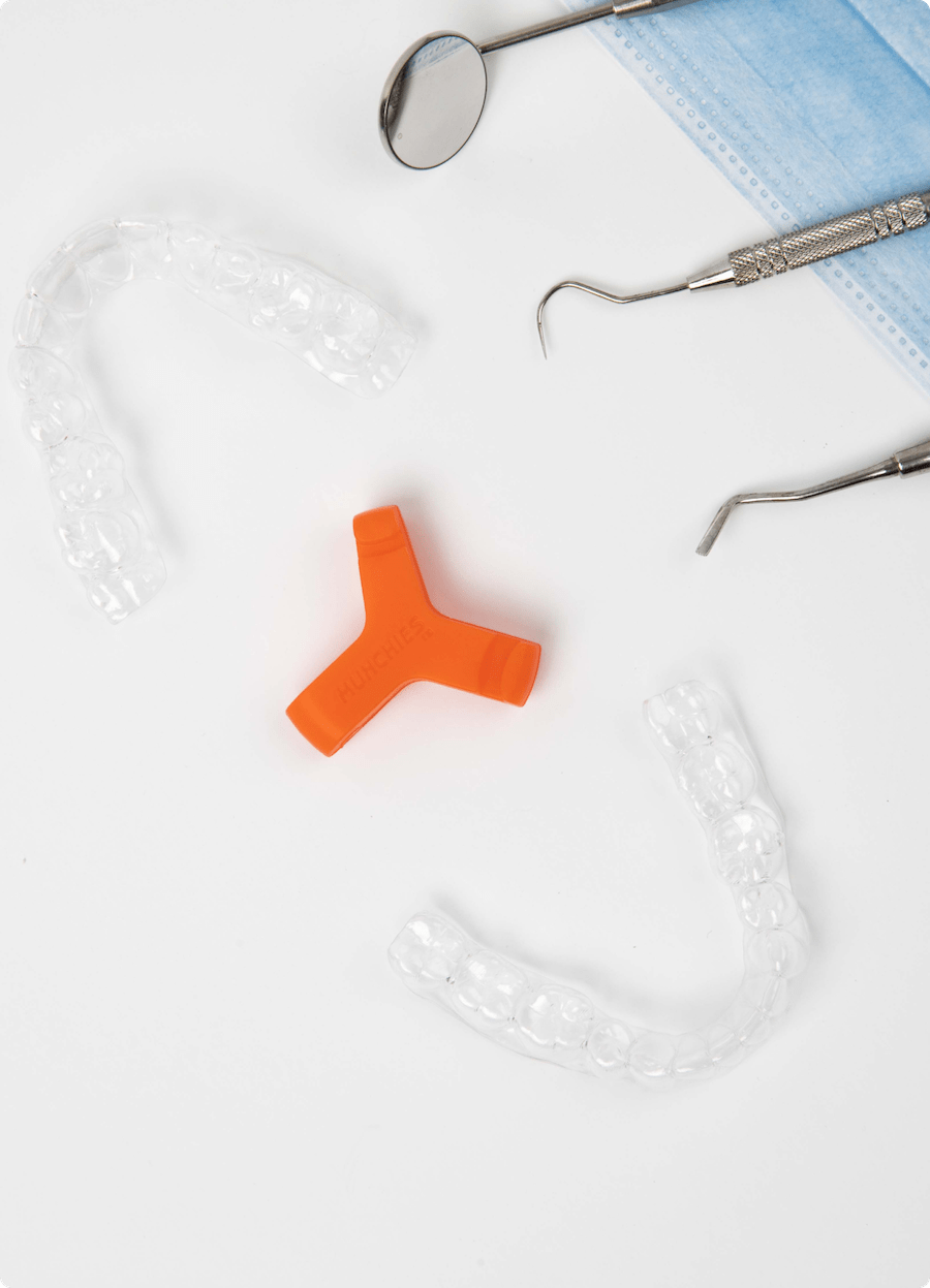 Orange Munchie surrounded by clear aligners and dental instruments laying on a face mask, against a white background